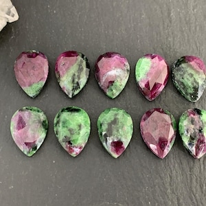 Op de afbeelding: Tien druppelvormige edelstenen met een mix van dieprode, groene en zwarte insluitsels. De edelstenen zijn gefacetteerd en vertonen een gepolijst oppervlak. De stenen zijn gerangschikt op een donkergrijze ondergrond, met een witte kristal op de achtergrond.
