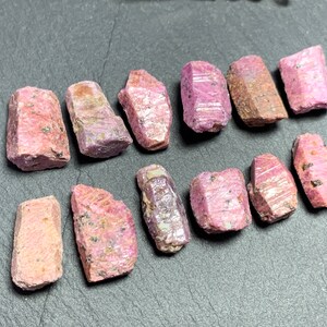 May include: A collection of 12 pink ruby rough crystals. The crystals are various sizes and shapes, with some having a more pointed shape and others being more rounded. The crystals are all a light pink color and have a shiny, translucent appearance.