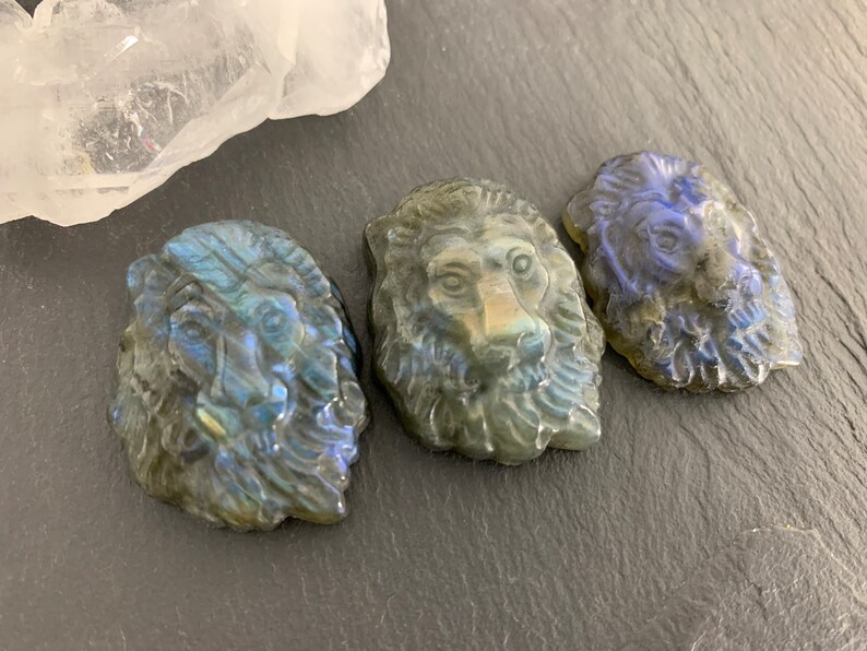 May include: Three carved labradorite lion head pendants. The pendants are a dark gray color with iridescent blue and green hues.