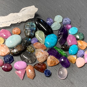 Könnte beinhalten: Eine Sammlung loser Edelsteine in verschiedenen Formen, Größen und Farben, darunter blaue, violette, rosa, orange, grüne und schwarze Steine. Ein Stein ist in Form eines Schädels geschliffen und ein klarer, länglicher Quarzkristall ist enthalten.