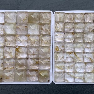 Pode incluir: Duas bandejas brancas cheias de pedras preciosas transparentes, cortadas em forma quadrada. Cada pedra tem inclusões douradas em forma de agulha. As pedras estão dispostas em um padrão de grade, mostrando sua clareza e estruturas internas únicas.