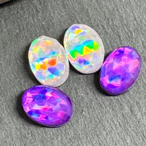 May include: Four oval-shaped gemstones with faceted surfaces. Two are iridescent with rainbow hues, and two are shades of purple. The gemstones are arranged on a textured gray surface.