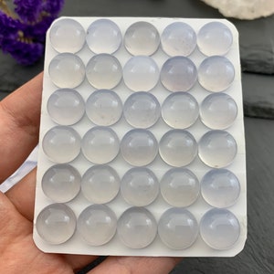 Op de afbeelding: Een verzameling van vijfendertig lichtblauwe, ronde cabochons, gerangschikt in een rasterpatroon op een witte kaart. De cabochons hebben een glad, gepolijst oppervlak. De achtergrond omvat een hand, paarse bloemen en een donker oppervlak.