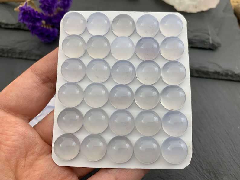 Op de afbeelding: Een verzameling van vijfendertig lichtblauwe, ronde cabochons, gerangschikt in een rasterpatroon op een witte kaart. De cabochons hebben een glad, gepolijst oppervlak. De achtergrond omvat een hand, paarse bloemen en een donker oppervlak.