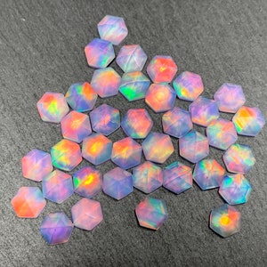 May include: A collection of iridescent, hexagonal gemstones. The gems display a range of colors, including pink, orange, blue, and green, with a shimmering, opalescent effect. The faceted stones are scattered on a dark gray surface.