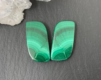 May include: Two polished malachite stones with a vibrant green color and unique banded patterns. The stones have a smooth, curved shape and are displayed on a dark gray surface. A cluster of white crystals is visible in the background.