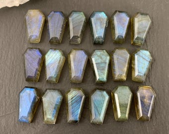 Op de afbeelding: Achttien gefacetteerde labradoriet edelstenen in de vorm van een doodskist. Elke steen heeft een gepolijst oppervlak met iriserende flitsen van blauw, goud en groen licht die reflecteren over de donkere, halfdoorzichtige minerale structuur.