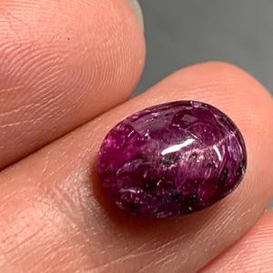 Pode incluir: Uma pedra preciosa de formato oval com corte cabochão e um tom magenta e roxo profundo. A superfície da pedra apresenta um acabamento polido e reflexivo com inclusões internas visíveis e manchas escuras em toda a sua estrutura.