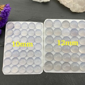 Op de afbeelding: Twee sets ronde, lichtblauwe edelstenen, &eacute;&eacute;n met het label "10mm" en de andere met "12mm". De edelstenen zijn in rijen op witte kaarten gerangschikt. Paarse gedroogde bloemen zijn linksboven zichtbaar.