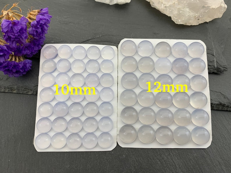 Op de afbeelding: Twee sets ronde, lichtblauwe edelstenen, &eacute;&eacute;n met het label "10mm" en de andere met "12mm". De edelstenen zijn in rijen op witte kaarten gerangschikt. Paarse gedroogde bloemen zijn linksboven zichtbaar.