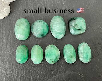May include: Ten faceted green emerald gemstones, each with a unique shape and cut, are arranged on a dark gray surface.  The stones are oval, round, and rectangular in shape.  The stones are all approximately the same size.