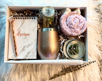 May include: A white gift box filled with various items, including a pink fuzzy slipper, a rose gold tumbler with the word "Girl" engraved on it, a small glass jar with a lid, a small notebook with the word "dream" written on it, a rose gold pen, and a small glass jar with a candle and a green succulent inside.