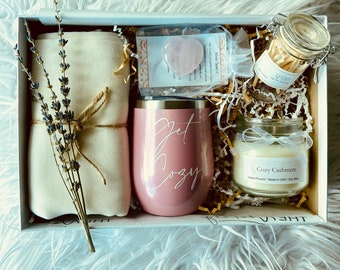 May include: A white gift box filled with a variety of items, including a pink tumbler with the words "Get Cozy" on it, a white candle with the text "Cozy Cashmere", a small jar of matches, a pink heart-shaped crystal, and a white cloth.