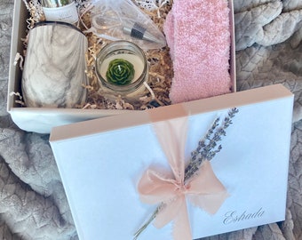 May include: A white gift box with a pink ribbon and lavender sprigs. The box is open and contains a white marble tumbler, a small glass jar with green contents, a candle with a green succulent inside, a clear plastic bag with a pair of scissors, a bottle stopper, and a pair of pink fuzzy socks. The box is labeled "Estrada".