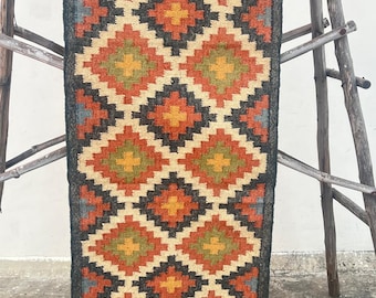 May include: A long, rectangular rug with a geometric pattern in shades of black, orange, brown, green, and cream. The rug has a fringe on the bottom edge.