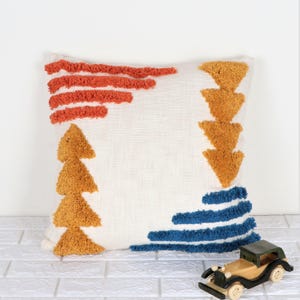 Puede incluir: Una almohada blanca con un diseño geométrico con detalles bordados en naranja, amarillo y azul. La almohada tiene un aspecto texturizado y esponjoso.