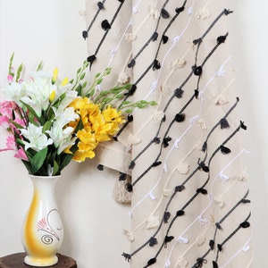 May include: A cream-colored throw blanket with black and white tassels. The blanket has a diagonal pattern of black and white stripes. The blanket is draped over a white wall and a wooden stool.