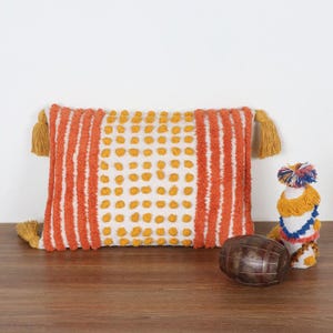May include: An orange and white rectangular pillow with yellow tassels on each end. The pillow has a striped design with a dotted pattern in the center. A decorative barrel and a small decorative item with tassels are also present.