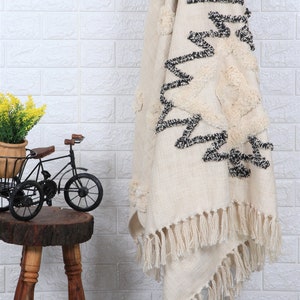 May include: A white throw blanket with black geometric designs and pom pom accents. The blanket has fringe on the bottom edge.