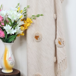 Può includere: Una coperta beige con un sottile motivo a spina di pesce e grandi pompon gialli e soffici. La coperta ha un bordo a frangia.