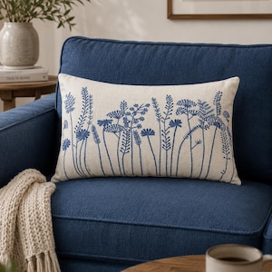 Può includere: Un cuscino rettangolare con una base in tessuto beige chiaro caratterizzato da motivi botanici ricamati in blu scuro di fiori di campo ed erbe. Il ricamo mostra varie altezze degli steli e forme floreali sulla superficie frontale.