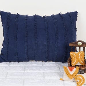 Puede incluir: Una almohada de tiro azul marino con un borde con flecos y una tela tejida texturizada. La almohada es rectangular y tiene un sutil patrón a rayas.