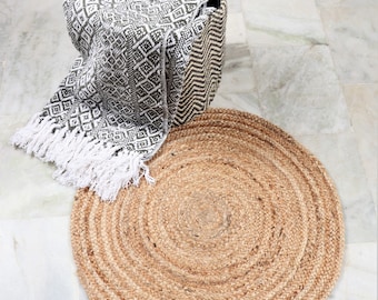 May include: A round, natural jute rug with a woven design. The rug has a light brown color and is placed on a white tiled floor. A black and white patterned blanket is in a woven basket.