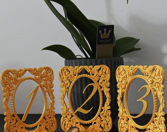 May include: Three gold-colored table number frames, each with a different number: 1, 2, and 3. The frames have an ornate, decorative design. A pair of glasses rests in front of the numbers. A potted plant and a tag that says "elegant" are in the background.