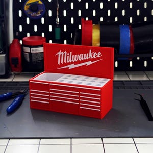 Puede incluir: Una caja de herramientas roja Milwaukee con letras blancas y un logotipo de rayo. La caja de herramientas tiene un compartimento superior y varios cajones. Unas pinzas azules y unas pinzas negras están sobre una alfombrilla gris delante de la caja.