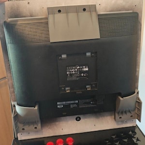 May include: The back of a black computer monitor mounted on a light-coloured panel with grey plastic support brackets. A central square section features a black label with white regulatory text, icons, and symbols, including CE and FCC logos.