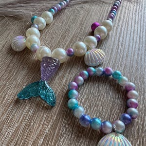 Könnte beinhalten: Ein Schmuckset zum Thema Meerjungfrau mit Halskette und Armband. Die Halskette hat große weiße Perlen, kleinere lila und blaue Perlen und einen Anhänger in Form einer Meerjungfrauenflosse. Das Armband hat passende Perlen und einen Muschel-Charm.