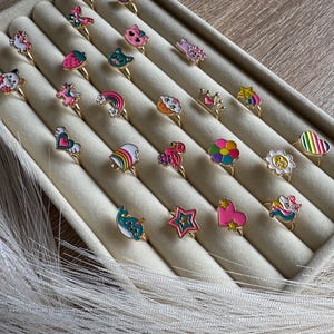 Könnte beinhalten: Eine Auswahl von 20 bunten Ringen mit verschiedenen Designs, darunter ein Einhorn, ein Regenbogen, ein Herz, ein Stern, ein Cupcake, ein Flamingo, eine Blume, ein Narwal, eine Katze, eine Erdbeere, ein Schmetterling, eine Krone, ein Smiley und eine Schleife. Die Ringe sind in Reihen auf einem beigefarbenen Samt-Hintergrund angeordnet.