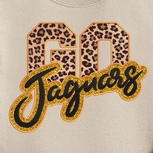 May include: A beige sweatshirt with the words "GO Jaguars" in a leopard print design and gold glitter lettering. The sweatshirt is a crew neck style and is made of a soft material. The design is centered on the front of the shirt.