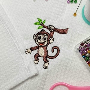 Puede incluir: Tela de tejido de gofre blanco bordada con un mono de dibujos animados colgado de una rama. El mono es marrón con una cara sonriente y hojas verdes. Suministros de costura, incluyendo alfileres e hilo, están esparcidos alrededor de la tela.