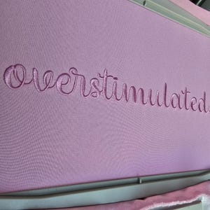 May include: A close-up of a pale purple fabric with the word "Overstimulated" embroidered in a matching pale purple thread. The text is in a cursive font. The fabric is stretched within an embroidery hoop.