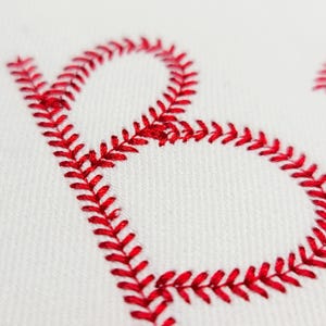 May include: Close-up of a white fabric with a red embroidered letter "B". The embroidery is in a baseball stitch pattern, with the red thread creating a bold, textured design. The background is a plain white fabric.