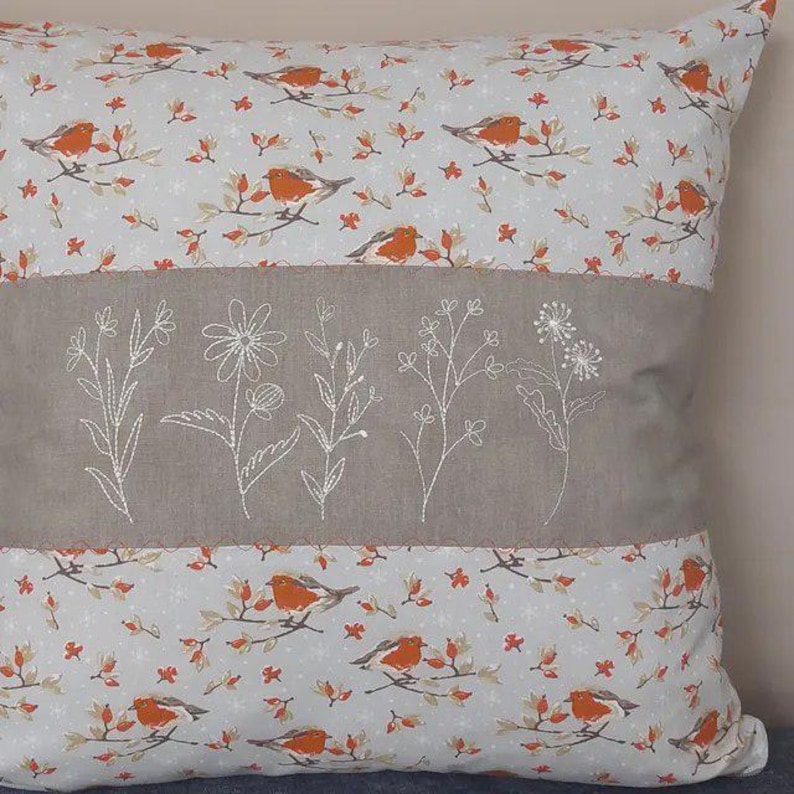 May include: Square decorative pillow with a light gray background and a pattern of orange birds on branches. A gray band with white embroidered flowers runs across the center. The pillow is a home decor item.