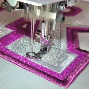 Può includere: Primo piano di un ago di macchina da cucire che ricama un disegno. Il disegno presenta un motivo geometrico a strati nei toni del rosa, argento e bianco. Il piedino e l'ago in metallo della macchina sono a fuoco, evidenziando il processo di ricamo.