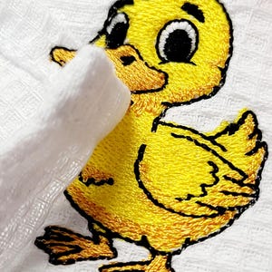 May include: Embroidered yellow duckling with black outlines on a white background. The duckling has a cheerful expression with large eyes and a small beak. The embroidery is detailed, and the duckling appears to be holding a white object.