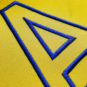 Puede incluir: Primer plano de una tela amarilla con una gran letra "A" bordada en azul. El bordado tiene un aspecto grueso y texturizado, y las puntadas están bien hechas. El fondo es de color amarillo liso.