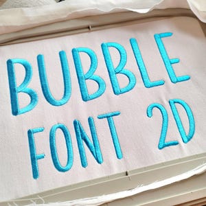 May include: Close-up of a white fabric with the words "BUBBLE FONT 2D" embroidered in bright turquoise thread. The letters are in a rounded, bubble-like font style, creating a playful and eye-catching design. The fabric is stretched within an embroidery hoop.