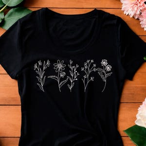 May include: Black t-shirt featuring a white embroidered floral design. The design includes various stylized flowers and stems. The shirt is made of a soft material and has a crew neck.