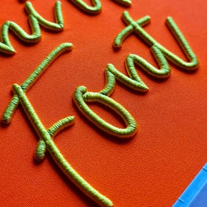 Puede incluir: Primer plano de texto bordado en cursiva. El texto está cosido con hilo dorado metálico sobre una tela naranja vibrante. La costura tiene una apariencia en relieve y texturizada, creando un efecto 3D. La tela tiene una textura sutil y tejida.