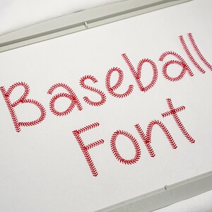 May include: Close-up of the words "Baseball Font" embroidered in red thread on white fabric. The letters are designed to resemble baseball stitching. The image is framed by a white embroidery hoop.