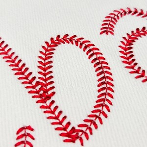 May include: Close-up of white fabric with the word "LOVE" embroidered in red baseball stitching. The letters are large and stylized, with the red thread creating a textured effect against the white background.