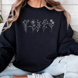 May include: A black sweatshirt featuring a delicate white floral line art design across the chest. The design includes various stylized flowers and stems. The sweatshirt is a crew neck style and appears to be made of a soft material.