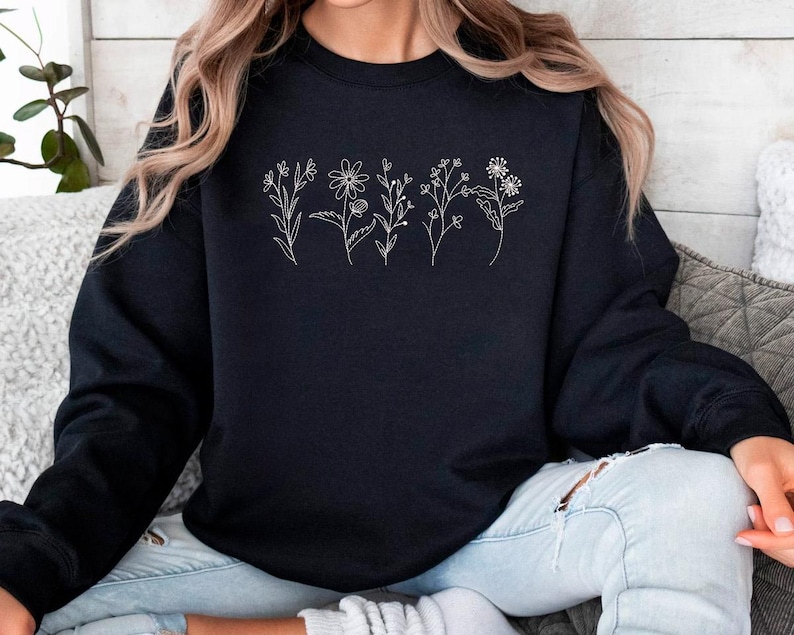 May include: A black sweatshirt featuring a delicate white floral line art design across the chest. The design includes various stylized flowers and stems. The sweatshirt is a crew neck style and appears to be made of a soft material.
