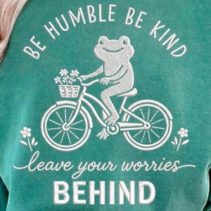 May include: Teal sweatshirt with a white graphic of a frog riding a bicycle, carrying flowers in a basket. The text "BE HUMBLE BE KIND" arches above the frog, with "leave your worries BEHIND" below.