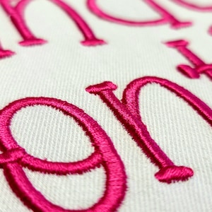 May include: Close-up of embroidered pink cursive letters on a white fabric background. The letters are densely stitched, creating a raised texture. The image highlights the detail of the embroidery.