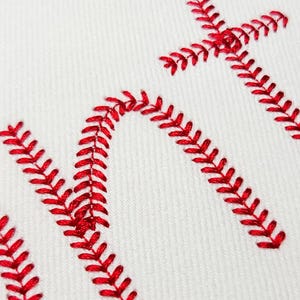 May include: Close-up of white fabric with red baseball-style stitching forming the letters "NT". The stitching is thick and detailed, creating a textured effect against the smooth white background. The design is likely for sports-themed apparel or decor.